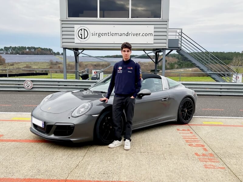 Jeune homme debout à côté d'une voiture de sport grise sur une piste de course, devant un bâtiment avec une enseigne.