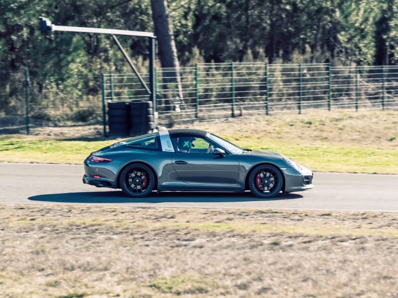Voiture de sport grise roulant sur une piste de course entourée d'herbe et de barrières de sécurité sous un ciel clair.