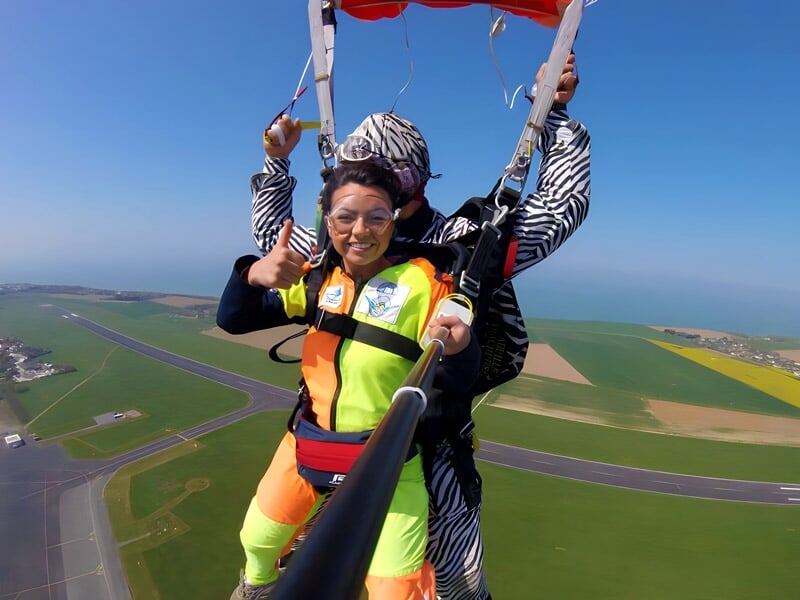 Un tandem de parachutistes descend en parachute au-dessus d'une piste d'aéroport avec un selfie stick en main.