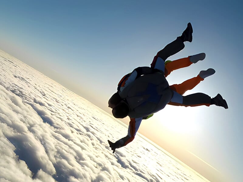 Deux parachutistes en chute libre au-dessus des nuages avec le soleil à l'horizon, en position horizontale.