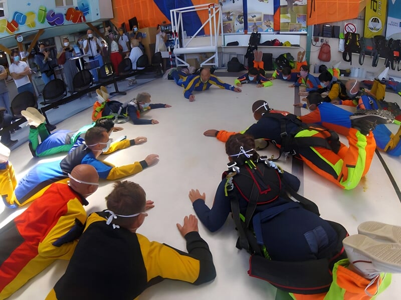 Groupe de parachutistes en combinaison colorée s'entraînant au sol dans un hangar avant leur saut.