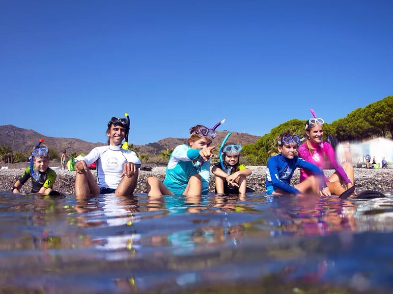 Un groupe d'enfants avec masques et tubas assis au bord de l'eau sur une plage ensoleillée.