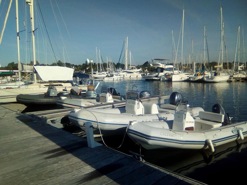 Bateaux et annexes amarrés à un ponton dans un port avec des voiliers en arrière-plan.