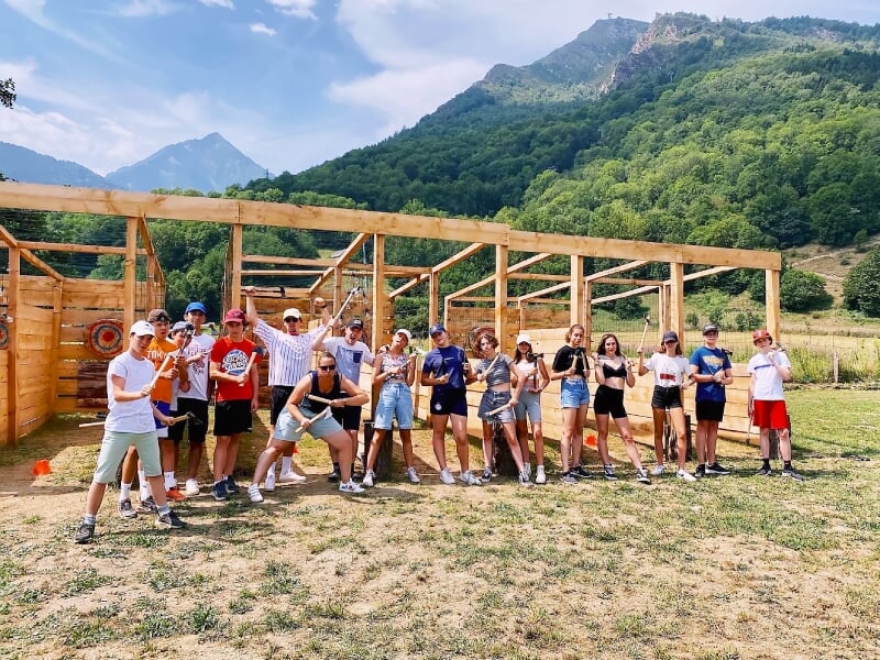 Groupe de personnes posant devant un terrain de lancer de haches en bois avec montagnes en arrière-plan.