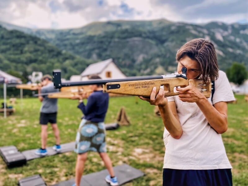 Jeune femme vise avec une carabine en bois, d'autres personnes s'entraînent au tir en arrière-plan en plein air.