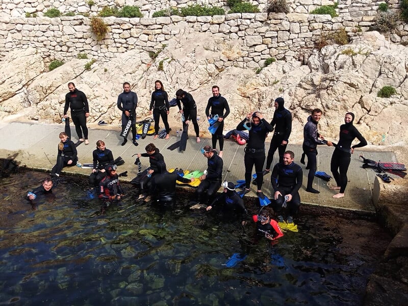 Groupe de personnes en combinaison de plongée au bord d'un bassin naturel entouré de rochers et d'un mur en pierre.