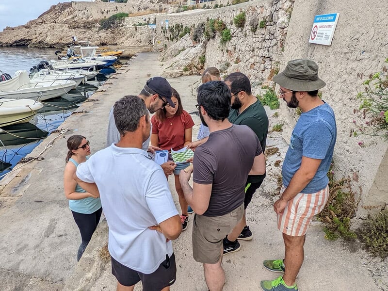 Groupe de personnes discutant sur un quai près de bateaux amarrés, avec un mur en béton et des rochers en arrière-plan.