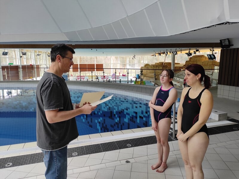 Un instructeur explique des consignes à deux jeunes filles en maillot de bain au bord d'une piscine intérieure.
