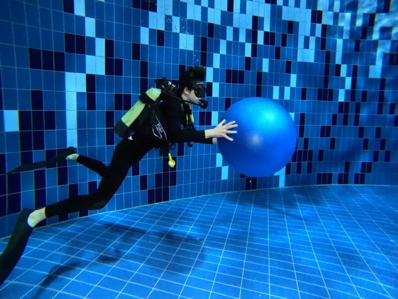 Un plongeur en combinaison complète pousse un gros ballon bleu sous l'eau dans une piscine aux carreaux bleus.