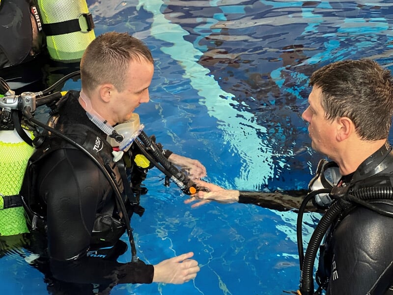 Deux plongeurs en combinaison noire s'entraînent dans une piscine, l'un tient un détendeur que l'autre examine.