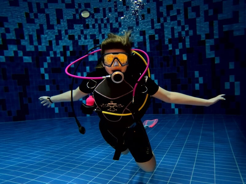 Une plongeuse en équipement complet flotte sous l'eau dans une piscine, bras étendus et masque de plongée.