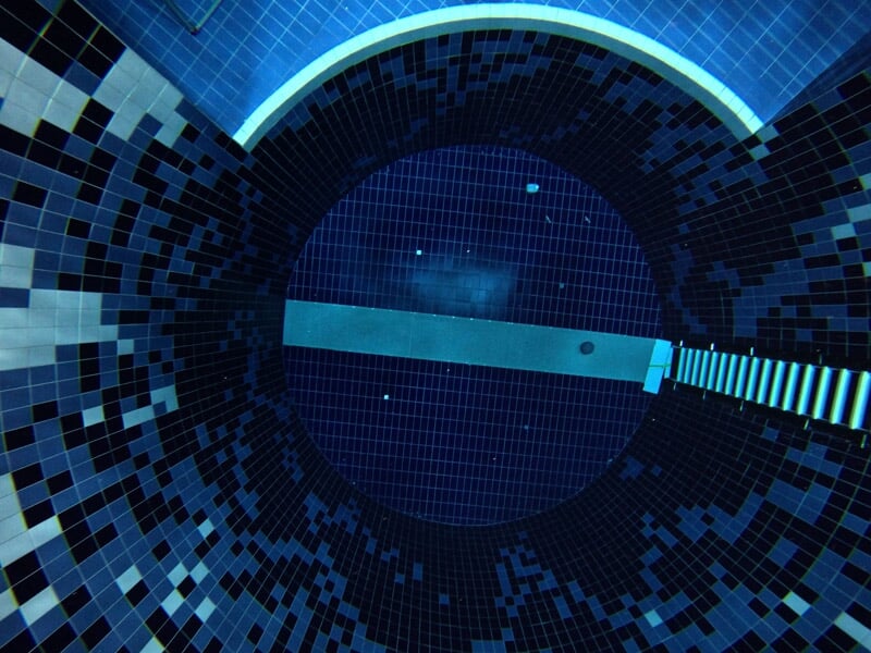 Vue plongeante d'une piscine circulaire avec un escalier et un tunnel, carrelage bleu et noir visible.