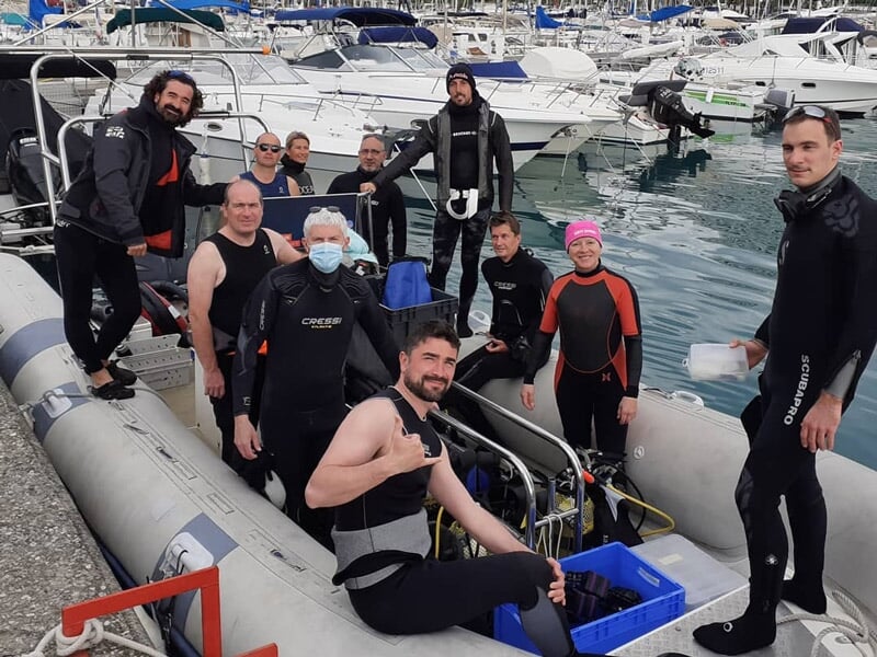 Groupe de plongeurs en combinaison noire posant sur un bateau pneumatique au port, prêts pour la plongée.