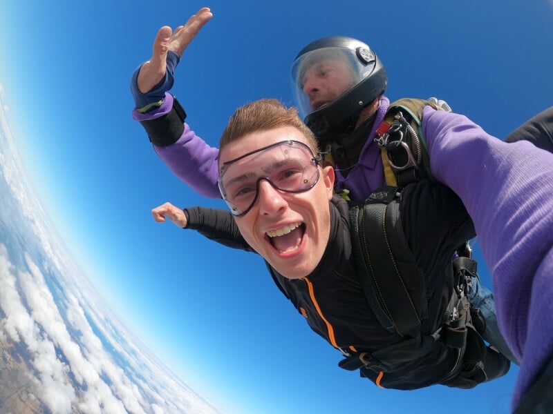 Un homme en parachute en chute libre sourit à la caméra, avec un ciel bleu et des nuages en arrière-plan.