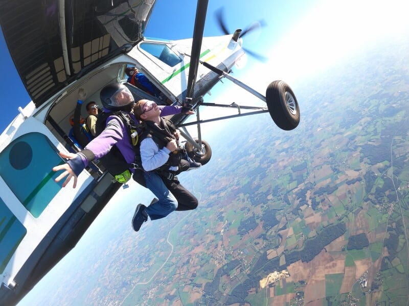 Deux parachutistes sortent d'un petit avion blanc en plein vol au-dessus d'un paysage rural.