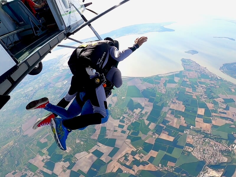 Un parachutiste en combinaison noire saute d'un avion au-dessus d'un paysage de champs et d'une ville.