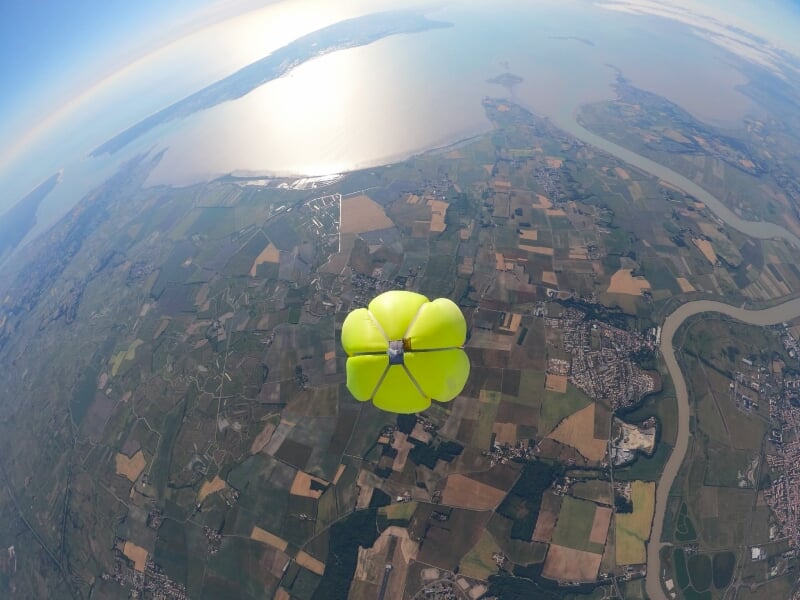 Parachute jaune ouvert en plein ciel au-dessus d'un paysage avec rivière, champs et villages visibles.