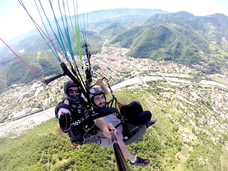 Deux personnes en parapente au-dessus d'une vallée verdoyante avec une ville en arrière-plan sous un ciel clair.