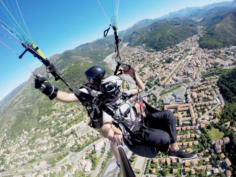 Vol en parapente en tandem au-dessus d'une ville entourée de collines et montagnes sous un ciel bleu lumineux.