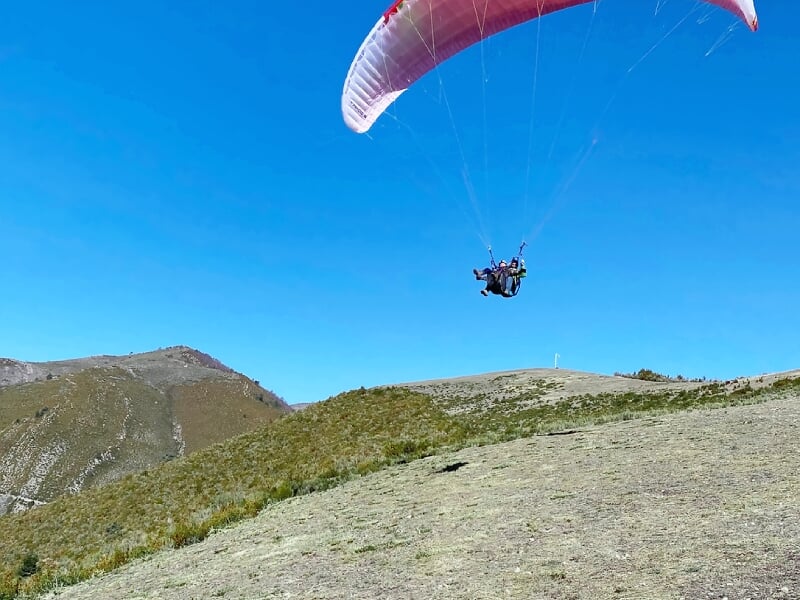 Parapente rouge en vol au-dessus d'un terrain montagneux avec des collines couvertes de végétation verte.