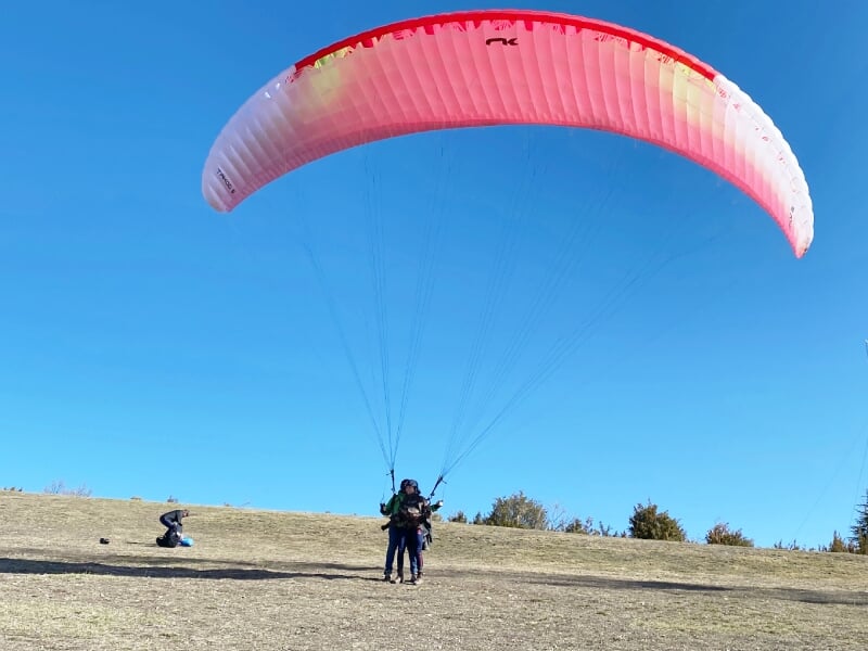 Départ au sol d'un vol en parapente avec deux personnes sous une voile rouge sur un terrain sec et dégagé.