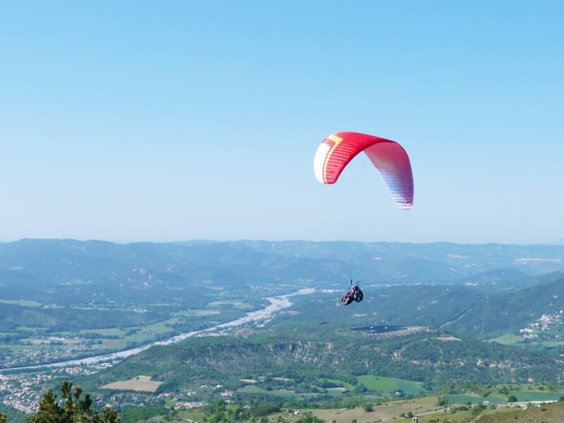 Parapente rouge en vol au-dessus d'une vallée avec une rivière et des collines verdoyantes sous un ciel dégagé.