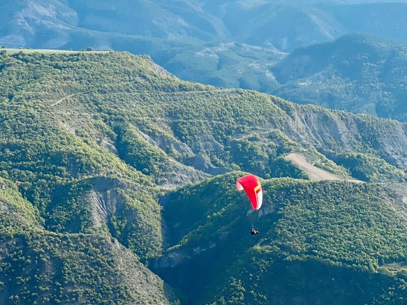 Parapente rouge survolant une zone montagneuse avec des vallées profondes et une végétation dense.