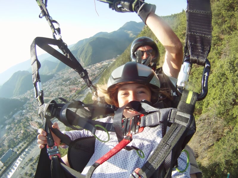 Deux personnes en parapente en tandem avec casque, souriantes, volant au-dessus d'une ville et des collines.
