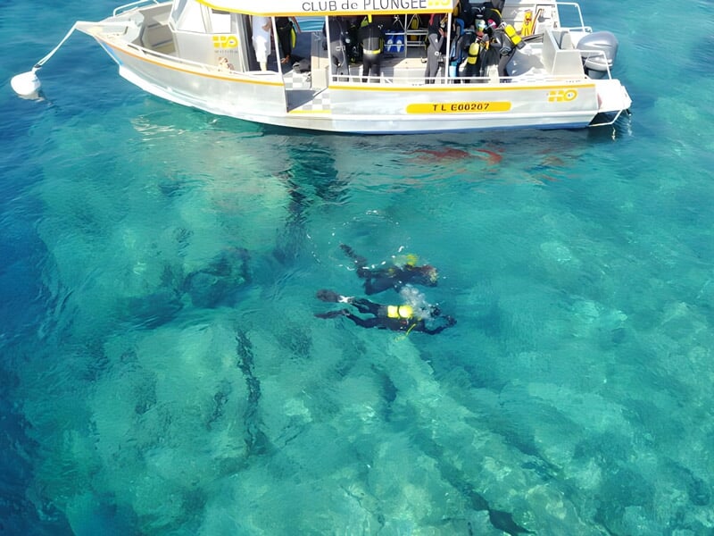 Deux plongeurs en combinaison noire avec bouteilles sous un bateau blanc sur une mer turquoise claire à Sainte-Maxime.