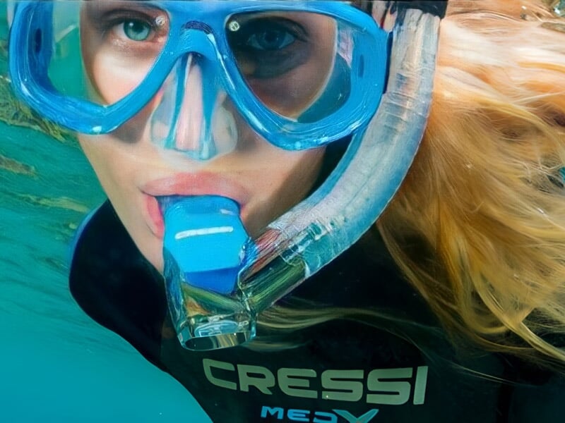 Plongeuse avec masque bleu et tuba, cheveux blonds flottant dans l'eau, regardant vers la caméra sous-marine.