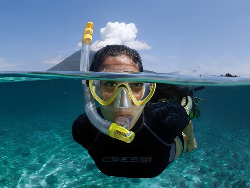 Plongeuse avec masque jaune et tuba, moitié sous l'eau, moitié hors de l'eau, dans une mer turquoise claire.