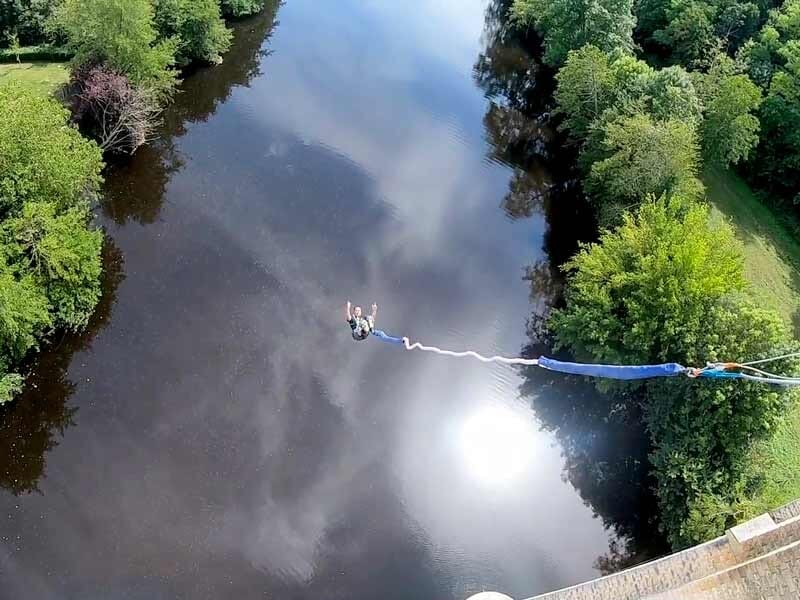 Vue plongeante d'une personne attachée à un élastique sautant dans le vide au-dessus d'une rivière entourée d'arbres.