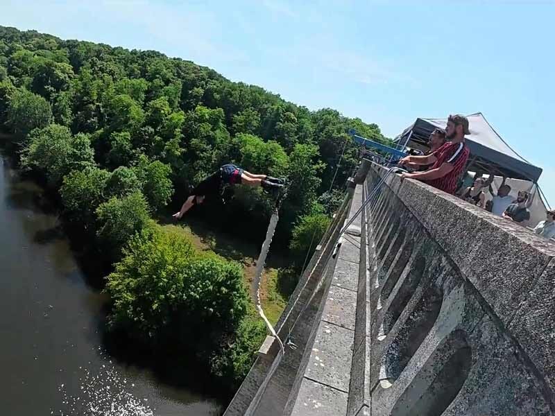 Saut à l'élastique depuis un pont avec spectateurs regardant, rivière et forêt en arrière-plan sous un ciel bleu.