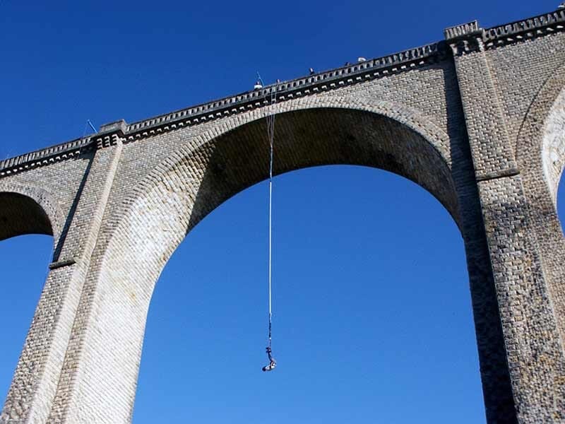 Personne suspendue à un élastique au milieu d'une arche en pierre du viaduc de Cluis sous un ciel bleu.