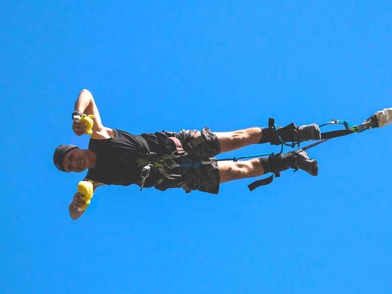 Homme souriant en plein saut à l'élastique, bras tendus, avec un ciel bleu en arrière-plan.