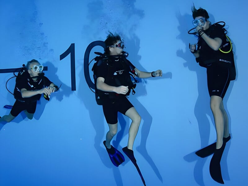 Trois personnes en tenue de plongée sous-marine dans une piscine, avec un mur marqué du chiffre 10 en arrière-plan.