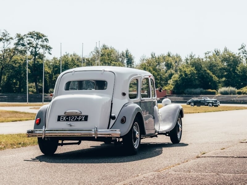 Voiture ancienne grise vue de dos roulant sur une route bordée d'arbres et d'espaces verts à Mulhouse.