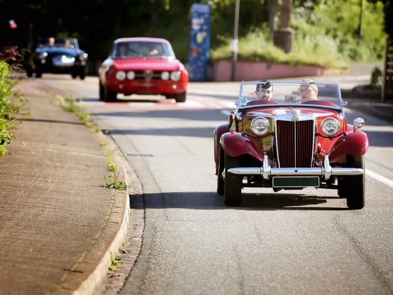 Voiture ancienne rouge conduite par deux personnes sur une route ensoleillée avec d'autres voitures en arrière-plan.