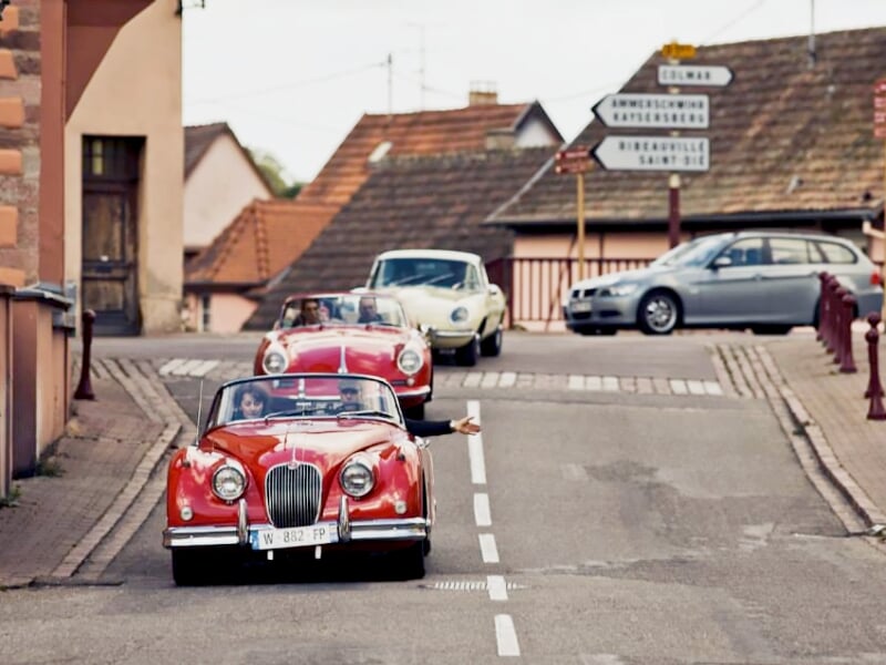 Voiture ancienne rouge décapotable roulant dans une rue de village avec plusieurs autres voitures et maisons.