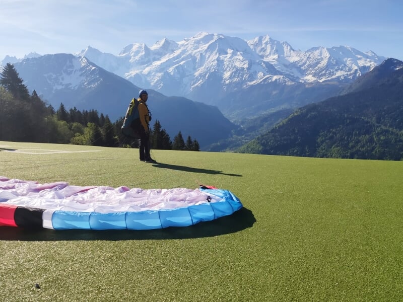 Parapente déployé au sol sur une aire d'herbe verte avec un homme debout face aux montagnes enneigées.