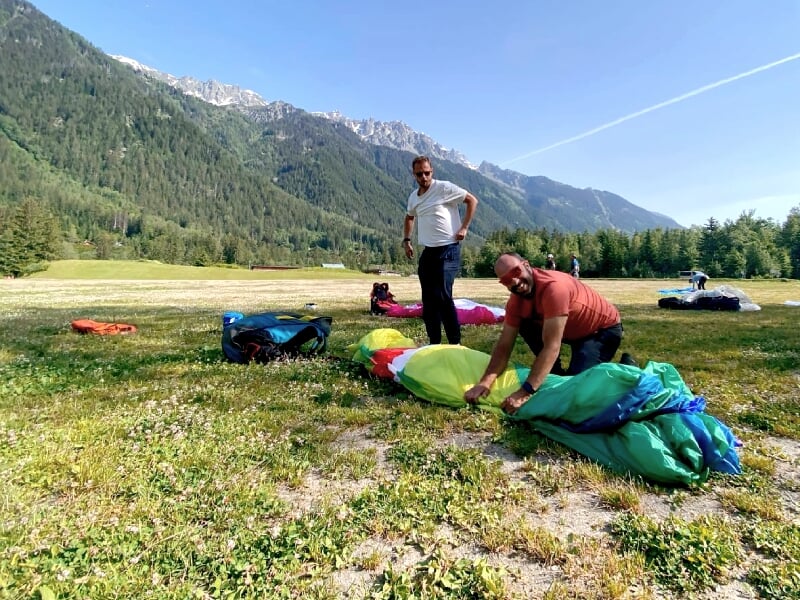 Deux hommes préparant leur équipement de parapente sur une prairie entourée de montagnes verdoyantes.