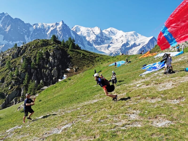 Personne en train de décoller en parapente sur une pente herbeuse avec montagnes enneigées en arrière-plan.