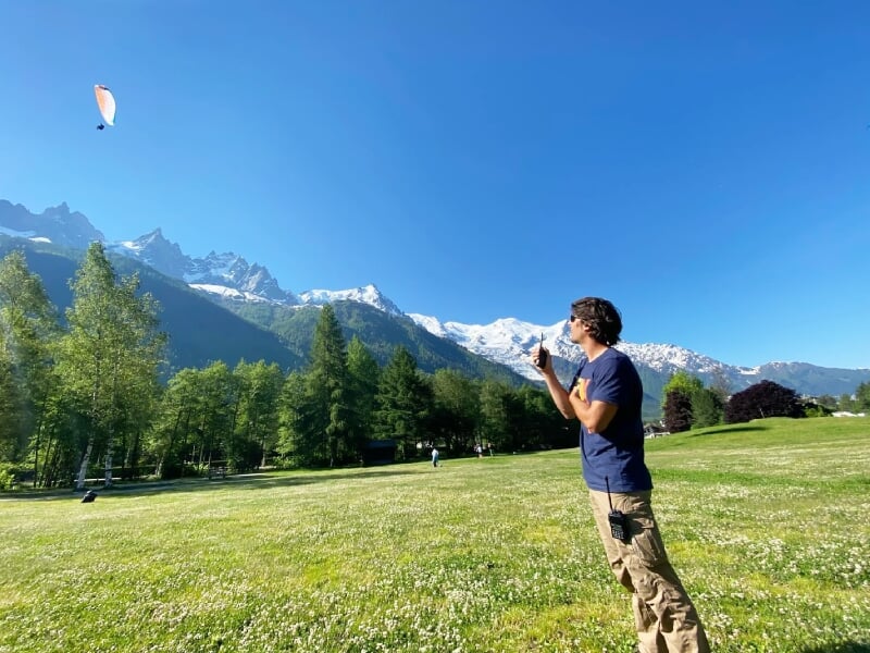 Homme observant un parapente en vol dans un champ vert avec montagnes enneigées et ciel bleu clair.