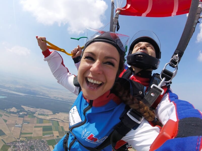 une femme sourit à la caméra lors d'un baptême de parachutisme avec un moniteur derrière elle