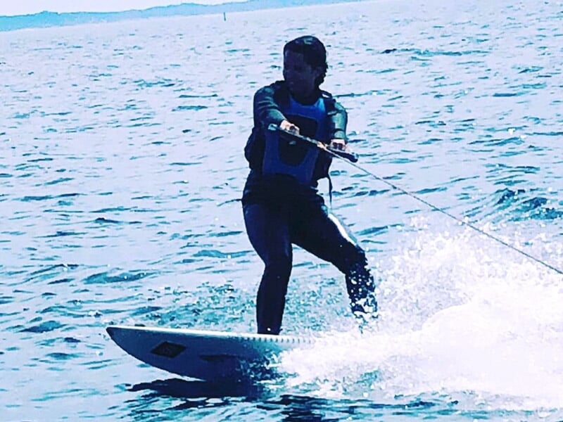 Personne en combinaison noire pratiquant le wakeboard sur une eau calme avec un ciel clair en arrière-plan.