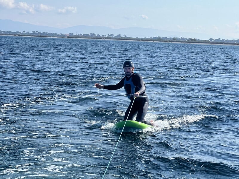 Homme en combinaison noire sur un wakeboard vert tiré par un bateau sur une étendue d'eau avec des montagnes au loin.