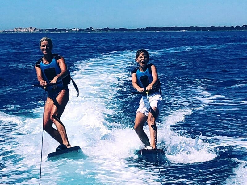 Deux personnes souriantes en gilets de sauvetage faisant du ski nautique côte à côte sur une mer bleue.