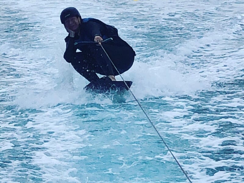 Homme en combinaison noire accroupi sur un wakeboard, glissant rapidement sur l'eau avec éclaboussures.