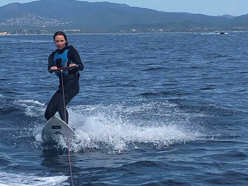 Femme en combinaison noire sur un wakeboard blanc, glissant sur une mer agitée avec des montagnes en arrière-plan.