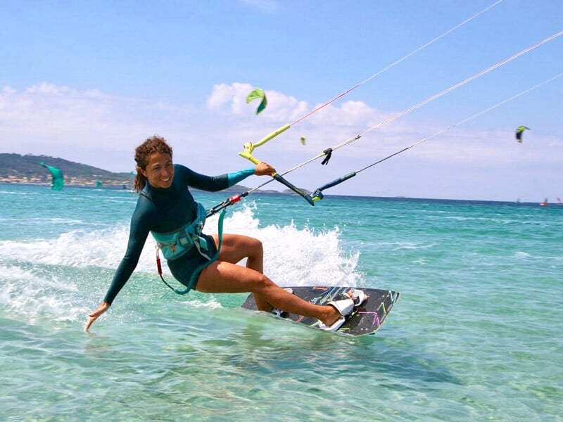 Femme en combinaison de kitesurf verte manœuvrant sa planche sur une eau turquoise claire sous un ciel ensoleillé.